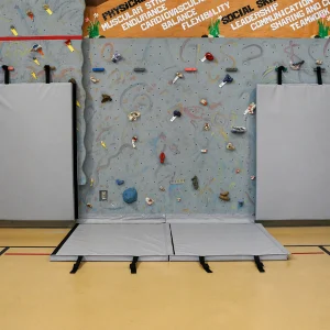 grey climbing wall mats in both the lowered and upright positions, protecting climbers from knocking into hand holds or landing on the hard gym floor surface